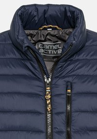 Marineblauwe pufferjas met een hoge kraag, een rits aan de voorkant en een gebrandmerkte binnenkant. Voorzien van horizontale stiksels en zwarte hardwareaccenten.