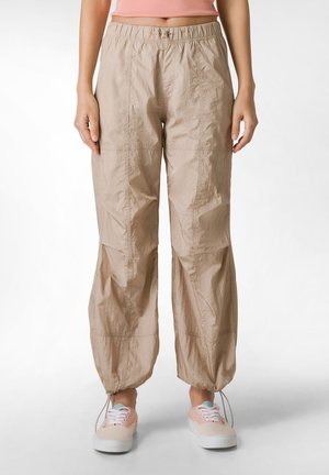 Lichtbeige, lichte stofbroeken met een elastische tailleband, taps toelopende manchetten en zichtbare stiksels. Gdragen met pastelgekleurde sneakers.