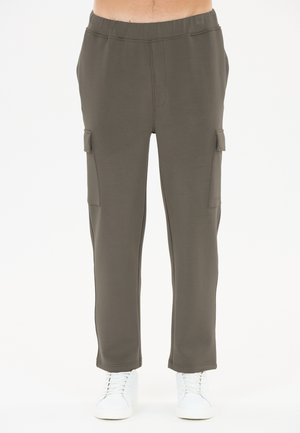 Man draagt olijfgroene cargobroek met elastische tailleband en witte sneakers, staand tegen een witte achtergrond.