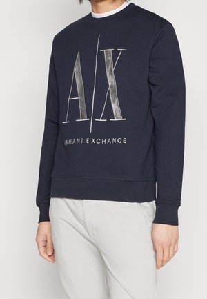 Man wearing navy blue sweatshirt with large "A|X" and "Armani Exchange" text, paired with light-colored pants against a plain background.