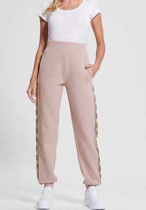 Kvinde iført beige joggebukser med sidelommer og hvide sneakers, kombineret med en enkel hvid kortærmet skjorte.