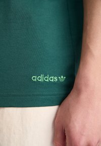 Green cotton t-shirt with a crew neck; features a stitched Adidas logo in light green on the bottom left. Smooth texture and fitted shape.