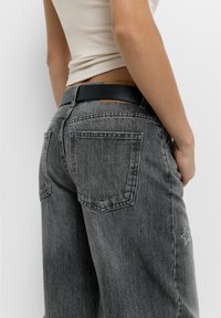 Szürke farmernadrág, laza illeszkedéssel, két hátsó zsebbel és egy fehér csillagmintával a jobb oldalon. Fekete öv tartozik hozzá.