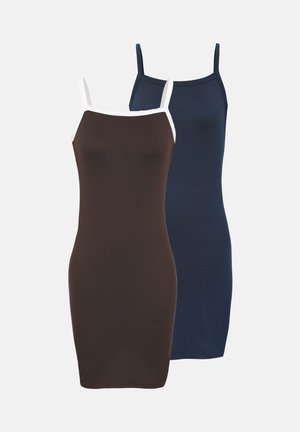 Kvinde med langt krøllet hår iført en tætsiddende brun kjole med hvide stropper og sorte plateau-sandaler, stående foran en ensfarvet hvid baggrund.