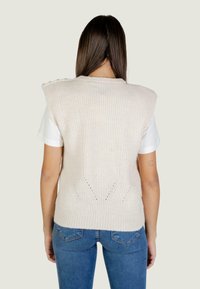 Gilet lavorato a maglia color beige chiaro con texture a coste, dettagli sulle spalle e scollatura a V sul retro. Abbinato a una camicia bianca e jeans blu.