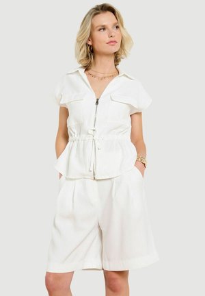 Blondínka v bielom krátkoteplom zipovacom topu s opaskom na stiahnutie v páse a širokých krátkych nohaviciach po kolená, ruky v kapse.