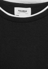 Lähikuva mustast kudumist sviitri kraest, millel on valge ääris ja Pull&Bear brändi silt krae sees.