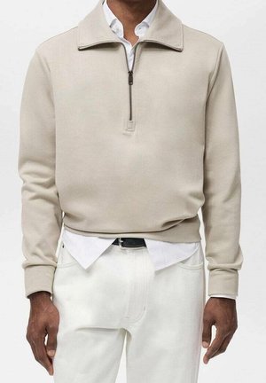 Mann trägt beigen Pullover mit Viertel-Reißverschluss über weißem Hemd mit Kragen, kombiniert mit weißen Hosen und schwarzem Gürtel, steht vor schlichtem Hintergrund.