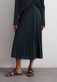 Una persona sta in piedi indossando una gonna plissettata scura e sandali neri, con un maglione scuro texture, sullo sfondo neutro.