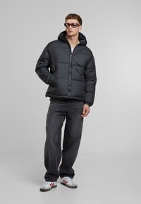 Sort oppustelig jakke med hætte, der har lynlås foran og sidelommer. Båret med mørkegrå jeans og hvide sneakers med røde detaljer.