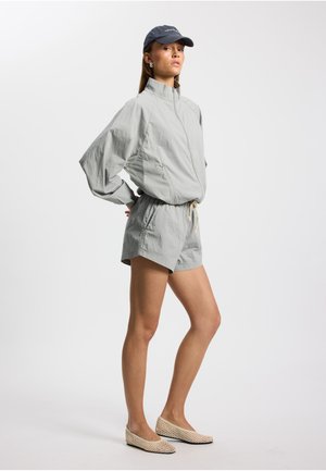 Lysegrå vindjakke med høy krage, kombinert med matchende shorts. Modellen har på seg beige flettede flate sko og en mørk caps, med hendene på hoftene.