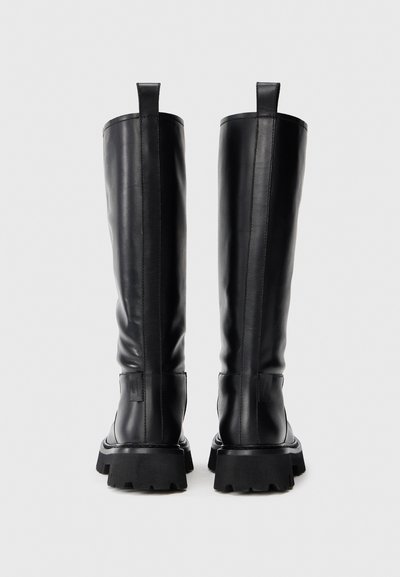 Bottes noires en cuir hautes avec des semelles à crampons épaisses et des languettes à l'arrière, vues de l'arrière sur un fond uni.