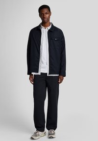 Marinblå jacka med en bröstficka, lagd över en vit randig skjorta, tillsammans med svarta byxor och grå sneakers.