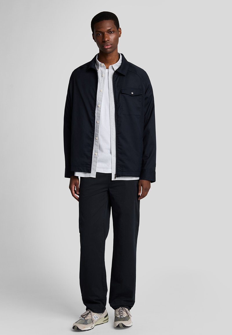 Marinblå jacka med en bröstficka, lagd över en vit randig skjorta, tillsammans med svarta byxor och grå sneakers.