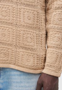 Jersey de punto beige con patrones cuadrados texturizados, usado con vaqueros de mezclilla azul claro, mostrando una mano de piel oscura descansando al lado.