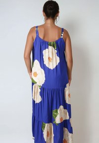 Blauwe bloemen maxi-jurk met grote crèmekleurige en groene bloempatronen, verstelbare schouderbanden met knopen, gerimpelde lagen en een gladde stoftextuur.