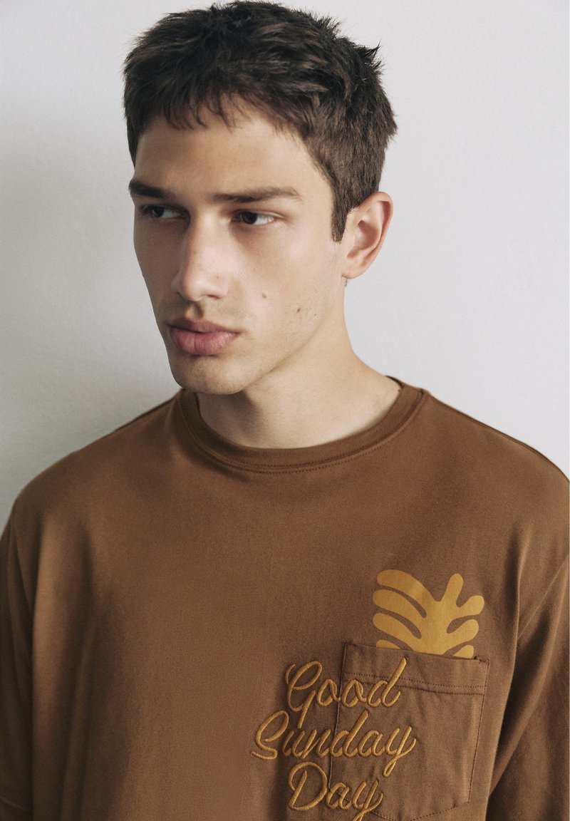 Jeune homme aux cheveux courts et foncés portant une chemise brune avec "Good Sunday Day" et un motif de feuille jaune au-dessus de la poche poitrine.
