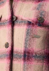 Lähedane vaade roosa ja beeži ruudulise villase jaki taskule, millel on tumedad nööbid ja metallist "Tommy Jeans" logo nõel.