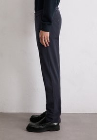 Marineblaue, schmal geschnittene Hosen aus Baumwollmischgewebe mit glatter Textur und seitlichen Taschen. Kombiniert mit schwarzen Slippers.