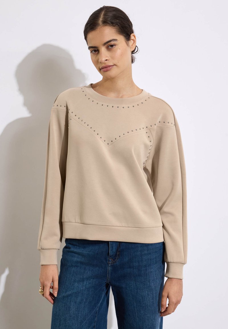 Felpa beige con maniche lunghe, scollatura rotonda, borchie decorative lungo le spalle e il petto, tessuto morbido, vestibilità regolare e orlo a coste.