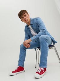 Chaqueta de denim azul clara sobre una camiseta blanca, combinada con jeans azul claros y zapatillas altas rojas. Modelo sentado en una silla.