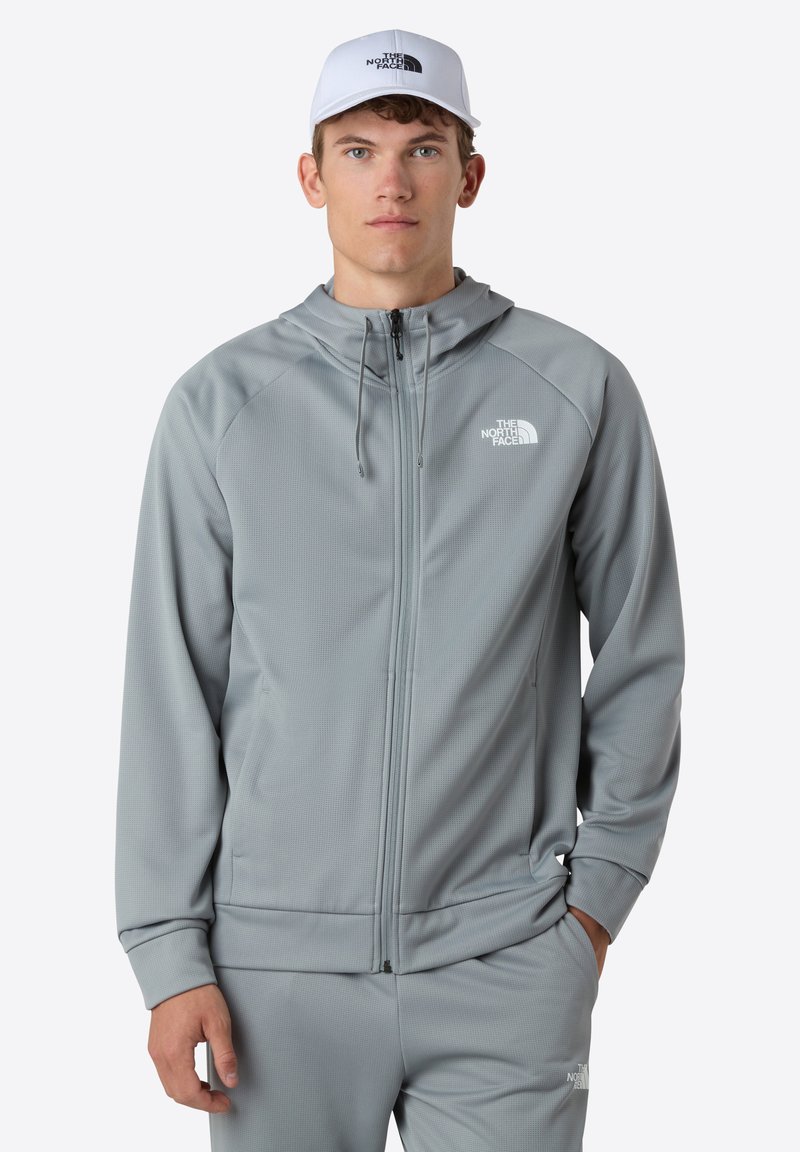 Grå zip-up hættejakke med en struktureret overflade, der har en hætte, sidelommer og The North Face-logoet i hvidt på venstre bryst.