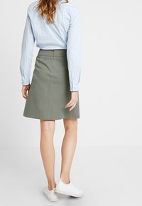 Una gonna in cotone verde oliva, lunga fino al ginocchio, con vita aderente e orlo svasato, abbinata a una camicia azzurro chiaro con bottoni e sneakers bianche.