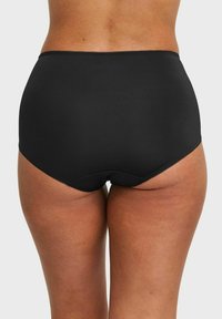 Vrouw in hoge zwarte onderbroek, zichtbaar van de taille tot halverwege de dijen, staande tegen een effen achtergrond.