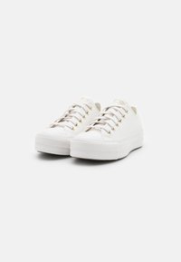Weiße Sneakers mit niedrigem Schnitt, weißen Schnürsenkeln und goldenen Ösen, mit dicken Gummisohlen, nebeneinander auf einem weißen Hintergrund platziert.