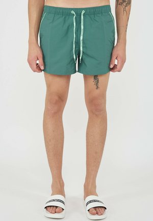 Man draagt groene zwembroek met trekkoord en witte Emporio Armani slippers, staand tegen een effen achtergrond.