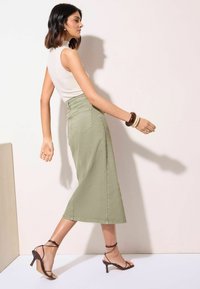 Olijfgroene denim midi-rok met een rechte snit, gecombineerd met een witte mouwloze top en zwarte gekruiste sandalen met hak.