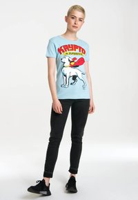 Light blue T-shirt featuring a graphic of a white dog with a red cape and yellow collar. Black skinny jeans and black sneakers are worn.