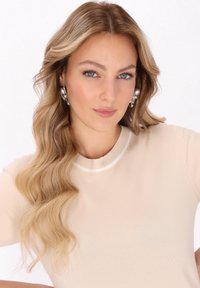 Blond włosy w delikatnych falach, nosząca beżowy sweter dziany z okrągłym dekoltem. Wyraźne duże srebrne i perłowe kolczyki. Jasne tło.