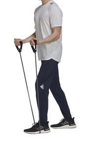 Person som bär en vit Adidas-skorta och mörkblå byxor använder motståndsband under fötterna för armträning.