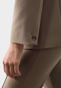 Primer plano de una mano junto a la manga de un traje beige con un botón y pantalones ajustados a juego.