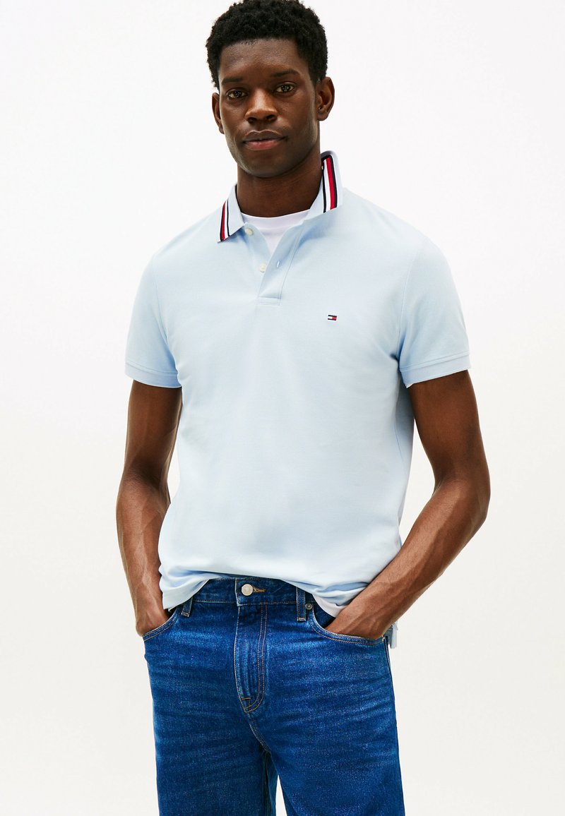 Polo shirt bleu clair avec un col rayé comportant des accents rouges et marins, porté avec un jean bleu foncé. La chemise a un petit logo sur la poitrine.