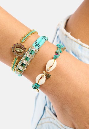 Quatre bracelets au poignet : tressé turquoise, perles dorées, orné de strass, et un avec des coquillages et des breloques. Mélange de matériaux et de textures.