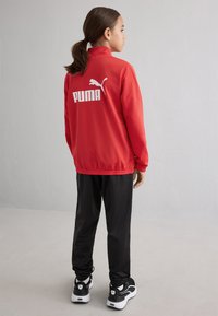 Crvena PUMA jakna s visokim ovratnikom i bijelim logotipom na leđima, uparena s crnim hlačama i crnim tenisicama s bijelim naglasima.