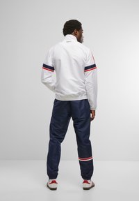 White windbreaker with red and navy stripes, elastic cuffs, paired with navy joggers featuring red accents, worn with white and red sneakers.