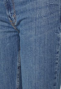Jeans de ganga azuis com um ligeiro desgaste, apresentando um design clássico de cinco bolsos, costura laranja e fecho de correr.