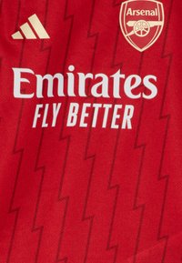 Camiseta deportiva roja con un patrón en zigzag, texto en blanco "Emirates FLY BETTER", logo dorado de Adidas y emblema del Arsenal en la esquina superior.