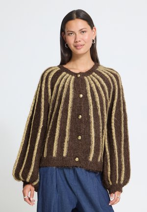Femme aux longs cheveux foncés porte un cardigan rayé marron et beige boutonné avec des manches bouffantes et un pantalon bleu, debout devant un fond uni.