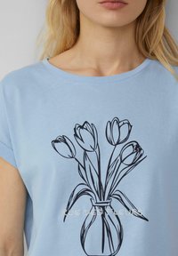 Hellblaues T-Shirt mit schwarzer Linienzeichnung von Tulpen in einer Vase. Weicher Stoff mit rundem Ausschnitt und kurzen, gerollten Ärmeln.