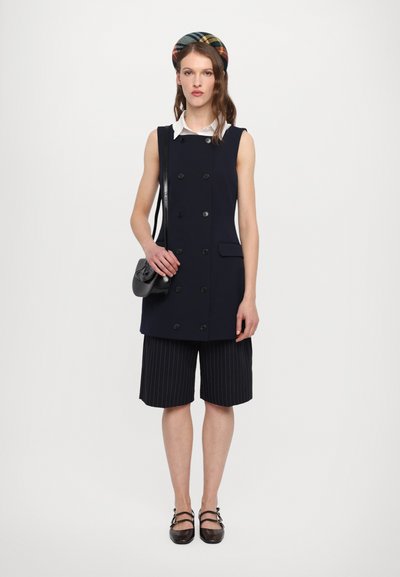 Vesta bleu în două rânduri de nasturi mari, asortată cu pantaloni scurți cu dungi. Accesoriile includ o bentiță cu model și o mică geantă neagră tip crossbody.
