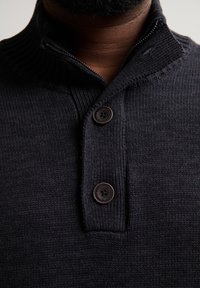 Dunkelblauer Strickpullover mit einem hohen Kragen, ausgestattet mit einem Reißverschluss und zwei braunen Knöpfen am Hals. Strukturstoff mit gerippten Details.