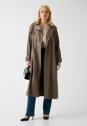 Femme blonde debout, portant un long trench-coat marron, un chemisier à volants, un jean bleu, des talons noirs, et tenant un petit sac à main noir.