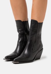 Stivali neri in pelle alla caviglia con punta affusolata, apertura scallopata, cuciture verticali e tacco a zeppa corto. Finiture in argento sulla punta.