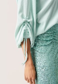 Blusa verde menta chiaro, textured con maniche arricciate e lacci, abbinata a una gonna teal scintillante con paillettes.