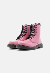 Ροζ δερμάτινα μποτάκια στρατιωτικού τύπου από λουστρίνι με μαύρα κορδόνια, κίτρινη ραφή και χοντρή σόλα από καουτσούκ. Ο σχεδιασμός περιλαμβάνει στρογγυλεμένη μύτη και ύψος μέχρι τον αστράγαλο.