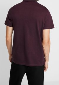 Homme portant un polo à manches courtes violet foncé et un pantalon noir, vu de dos contre un fond clair uni.